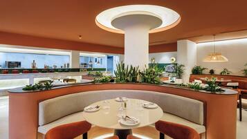 A modern restaurant interior with a central round table, cushioned seating, and a variety of plants.