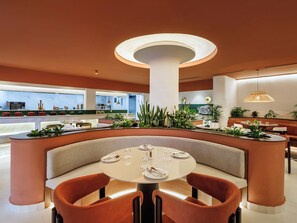 A modern restaurant interior with a central round table, cushioned seating, and a variety of plants. - Bagaincha Resort & Spa (Pokhara)