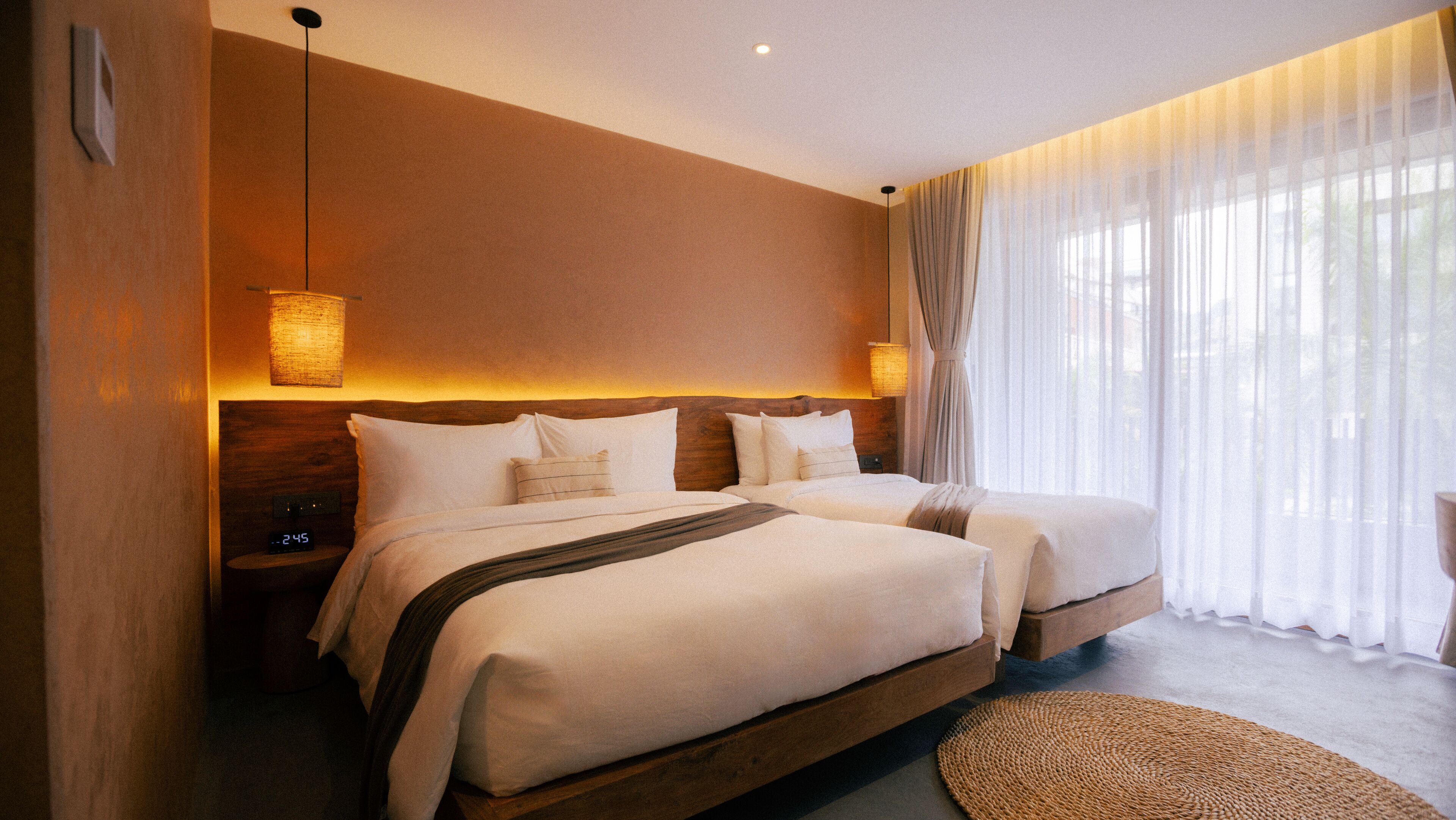 A hotel room with two beds, a large window with sheer curtains, and a woven rug on the floor.