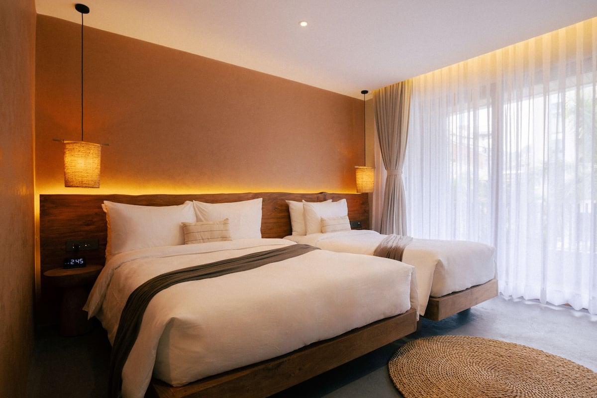 A hotel room with two beds, a large window with sheer curtains, and a woven rug on the floor.