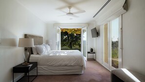 Un dormitorio con cama, mesitas de noche, ventilador de techo, televisor y un ventanal con vistas a árboles.
