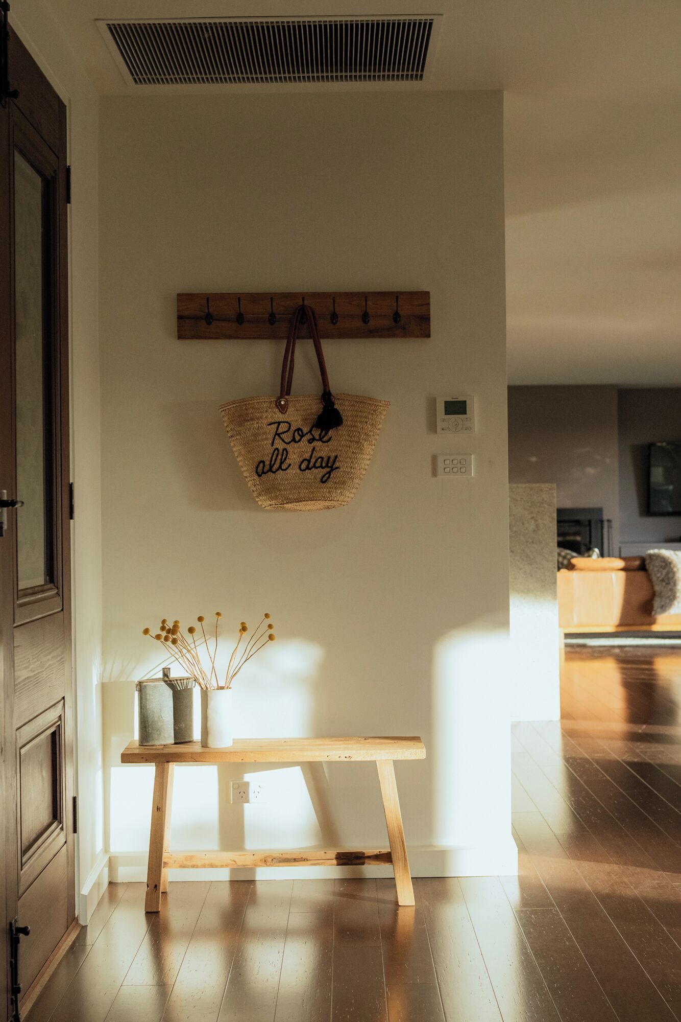 Un pasillo con un banco de madera, una cesta con asa fijada a la pared y una mesita con un florero de flores secas.