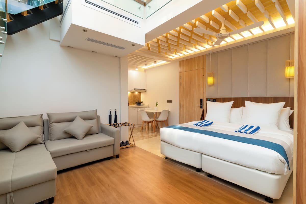 A modern hotel room with a large bed, a sofa, a desk, and a kitchenette.
