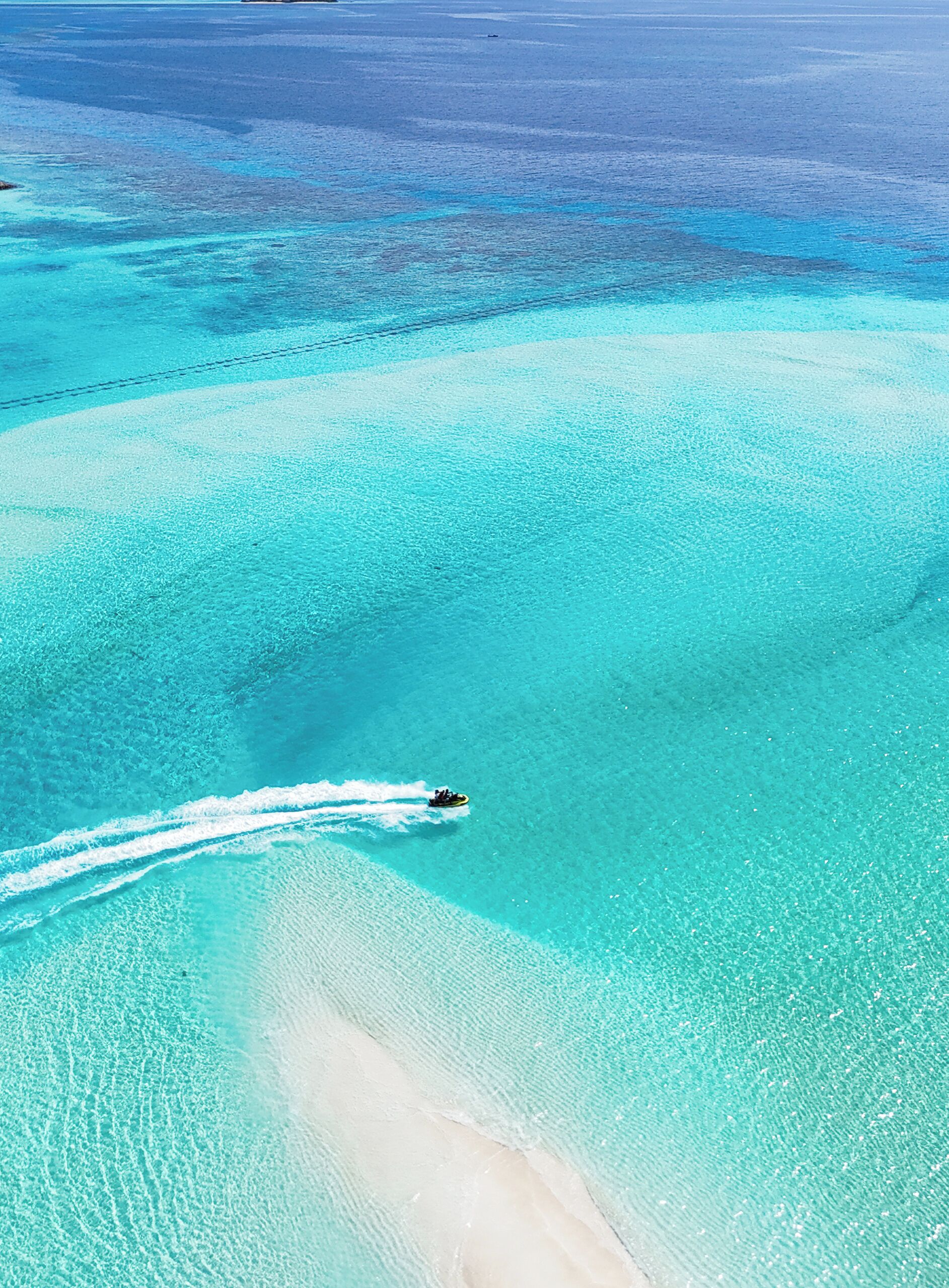 Une mer turquoise aux eaux limpides, traversée par un bateau laissant une vague derrière lui, bordée d’une plage de sable blanc et d’eaux peu profondes.