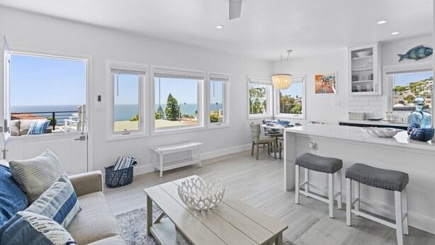 A spacious living area with a large window overlooking the sea, a dining area with a view, and a kitchen with white cabinetry. - Penthouse! Steps from Victoria Beach, 180 degree ocean views! (Laguna Beach)