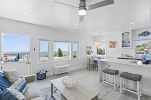 Een ruime woonkamer met een groot raam met uitzicht op zee, een eethoek met uitzicht en een keuken met witte kastjes.