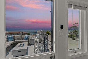 A balcony with a white sofa, a small table, and a white chair. The balcony overlooks a scenic view of the ocean and a sunset sky.