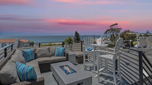 A rooftop terrace with a gray sofa, white chairs, and a small table with a drink. The terrace overlooks a scenic view of the ocean and a sunset.