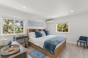 Un dormitorio con una cama de madera, dos ventanas con vistas a zonas verdes, una mesita de noche con una lámpara y una silla.