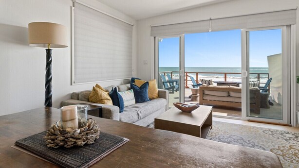 A modern living room with a sofa, coffee table, and a lamp. A large window offers a view of the beach and ocean. - Private beach, toes on the Sand!  Waterworld! STR23-0121 (Dana Point)