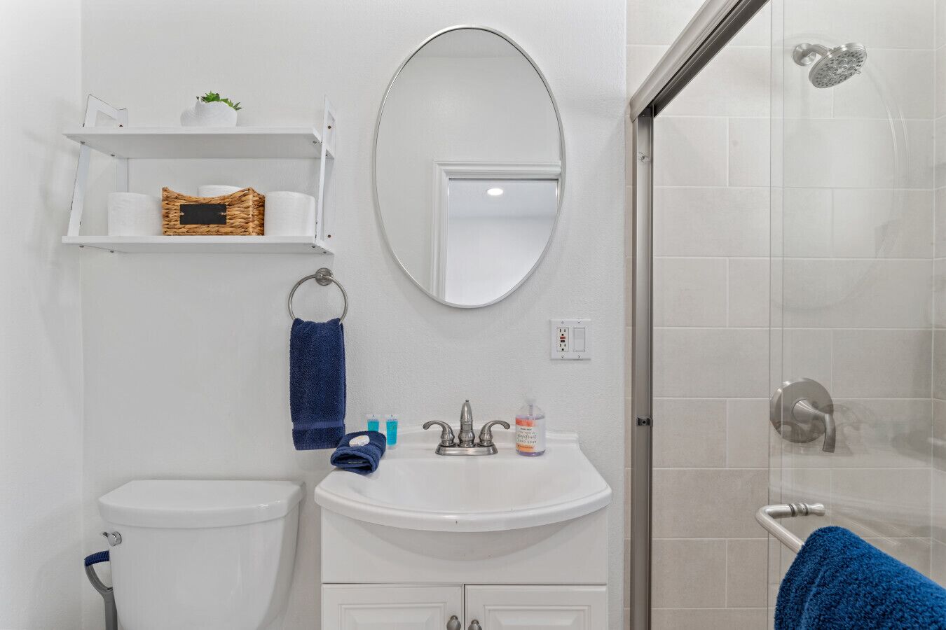 A bathroom with a toilet, sink, and shower. There is a towel rack with a blue towel, a mirror, and a shelf with toiletries.