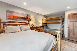 A bedroom with a large bed, a bunk bed, a wooden nightstand, a lamp, and a painting on the wall.