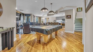 A spacious living room with a pool table, wooden flooring, and pendant lighting.