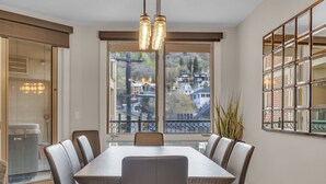 A dining area with a large table and six chairs, a view of a residential neighborhood through a large window, and a decorative light fixture above the table.