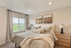 A bedroom with a large bed, two bedside tables with lamps, a window with curtains, and two landscape paintings on the wall.