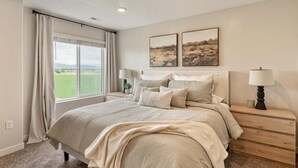 A bedroom with a large bed, two bedside tables with lamps, a window with curtains, and two landscape paintings on the wall.