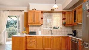 A kitchen with wooden cabinets, a built-in oven, a refrigerator, and a window with a view of greenery. - OLIA  APARTMENTS LYGIA (Lefkada)