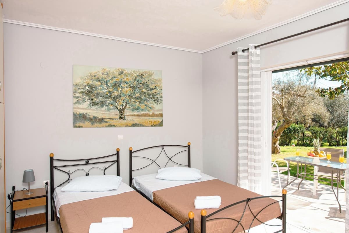 Ein Zimmer mit zwei Betten, einem Baumgemälde an der Wand und Blick auf einen Außenbereich mit Tisch und Früchten.