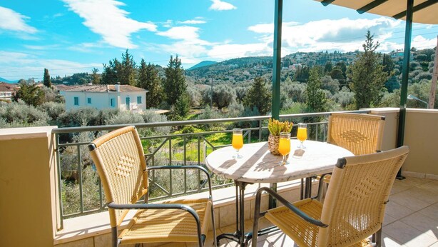 A balcony with wicker chairs and a table, overlooking a scenic landscape with trees and buildings. - OLIA  APARTMENTS LYGIA (Lefkada)