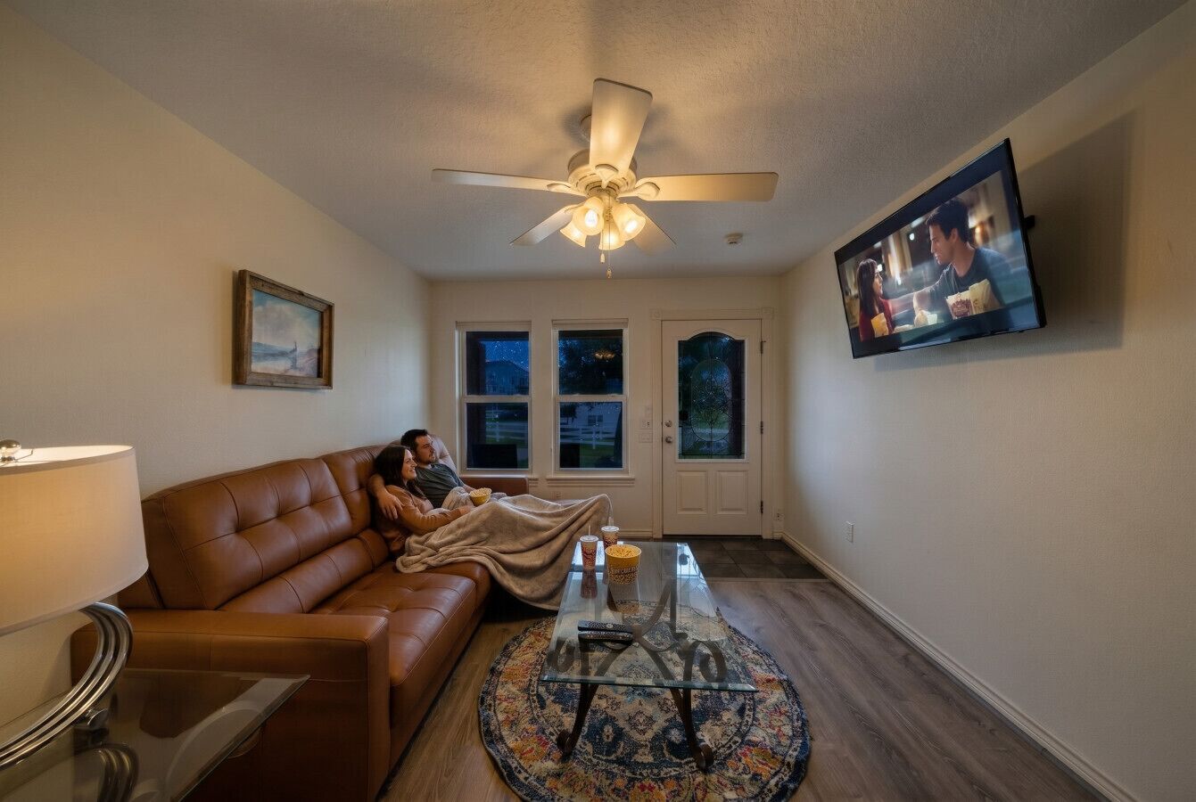 A living room with a brown leather sofa, a glass coffee table, a ceiling fan, a TV mounted on the wall, and a door with windows.