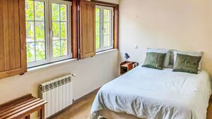 A bedroom with a bed, a nightstand, a window with curtains, a radiator, and a wooden bench.