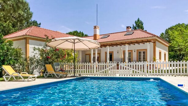 A swimming pool with a white fence, a covered patio with chairs, and a house with a red-tiled roof.
