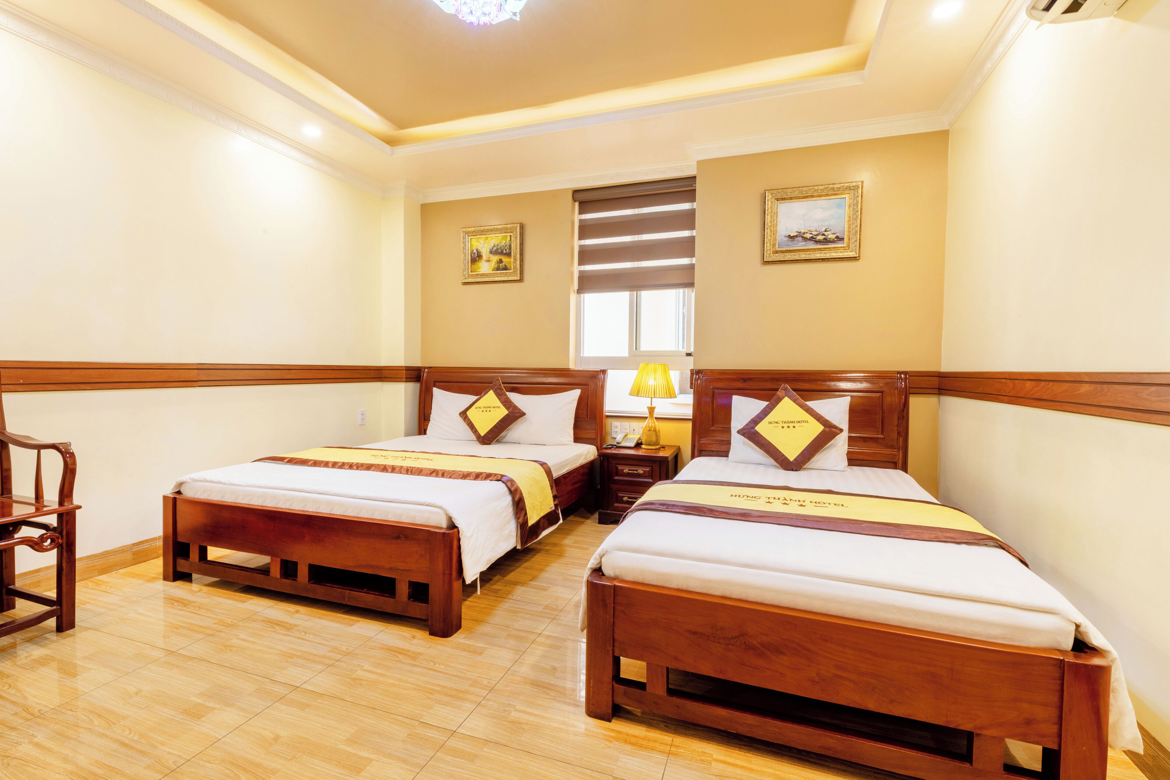 A hotel room with two wooden beds, a wooden chair, a nightstand with a lamp, and a window with blinds.