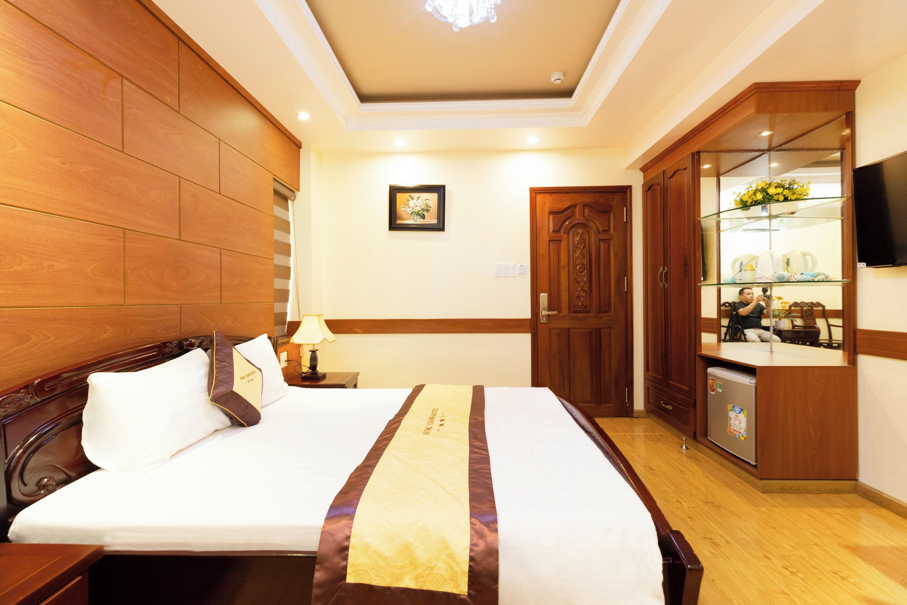 A hotel room with a large bed, wooden paneling on the walls, a small bedside table with a lamp, a wooden wardrobe, and a television on the wall.