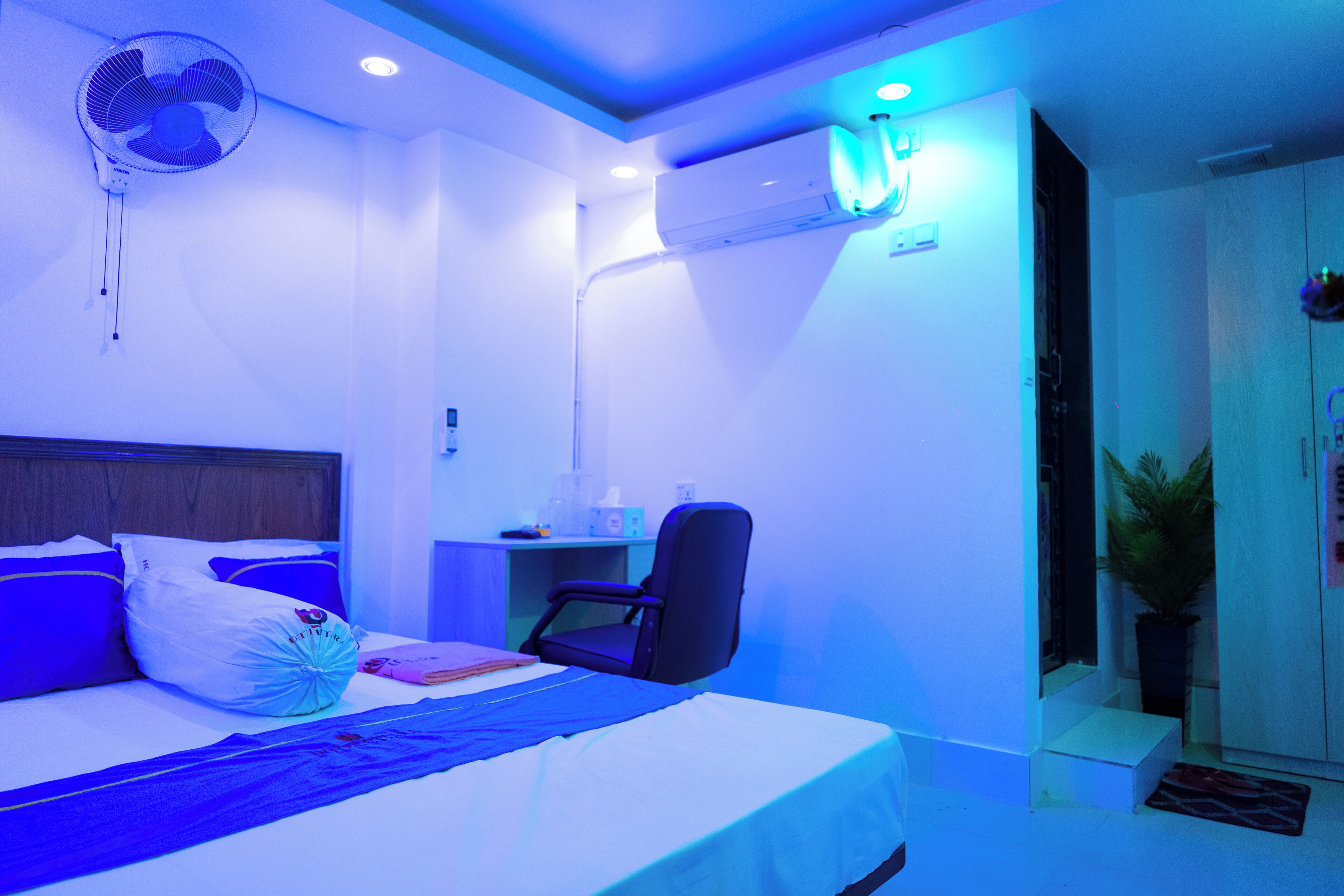 A hotel room with a bed, a desk, a chair, a fan, and an air conditioning unit.