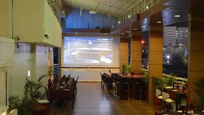 A spacious indoor area with a large screen displaying a presentation, multiple tables and chairs arranged for dining, and a view of the cityscape through large windows. - Hotel Somdeep Palace (Indore)