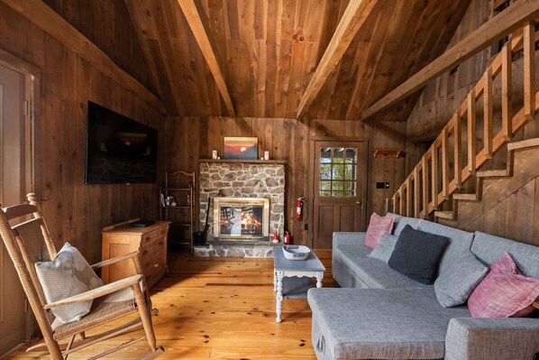 L’intérieur d’un chalet accueillant, avec une cheminée en pierre, des murs en bois, un fauteuil à bascule en bois, un canapé gris et un escalier.