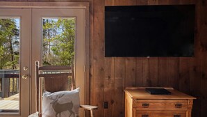 Una habitación con pared de madera, un televisor sobre un mueble de madera, una silla con cojín y una puerta que da al exterior.