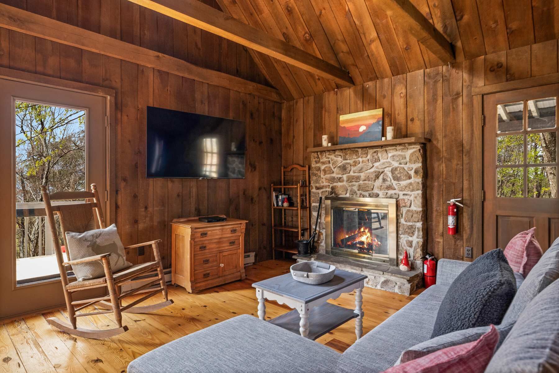 A cozy cabin interior with a stone fireplace, wooden walls, a television, and a rocking chair.