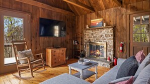 Interior acogedor de una cabaña con chimenea de piedra, paredes de madera, televisor y una mecedora.