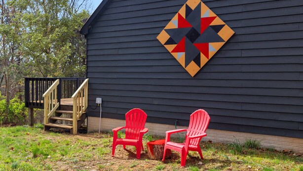 Una terraza de madera con dos sillas Adirondack rojas, una escalera de madera y un edificio negro con un adorno de pared geométrico.