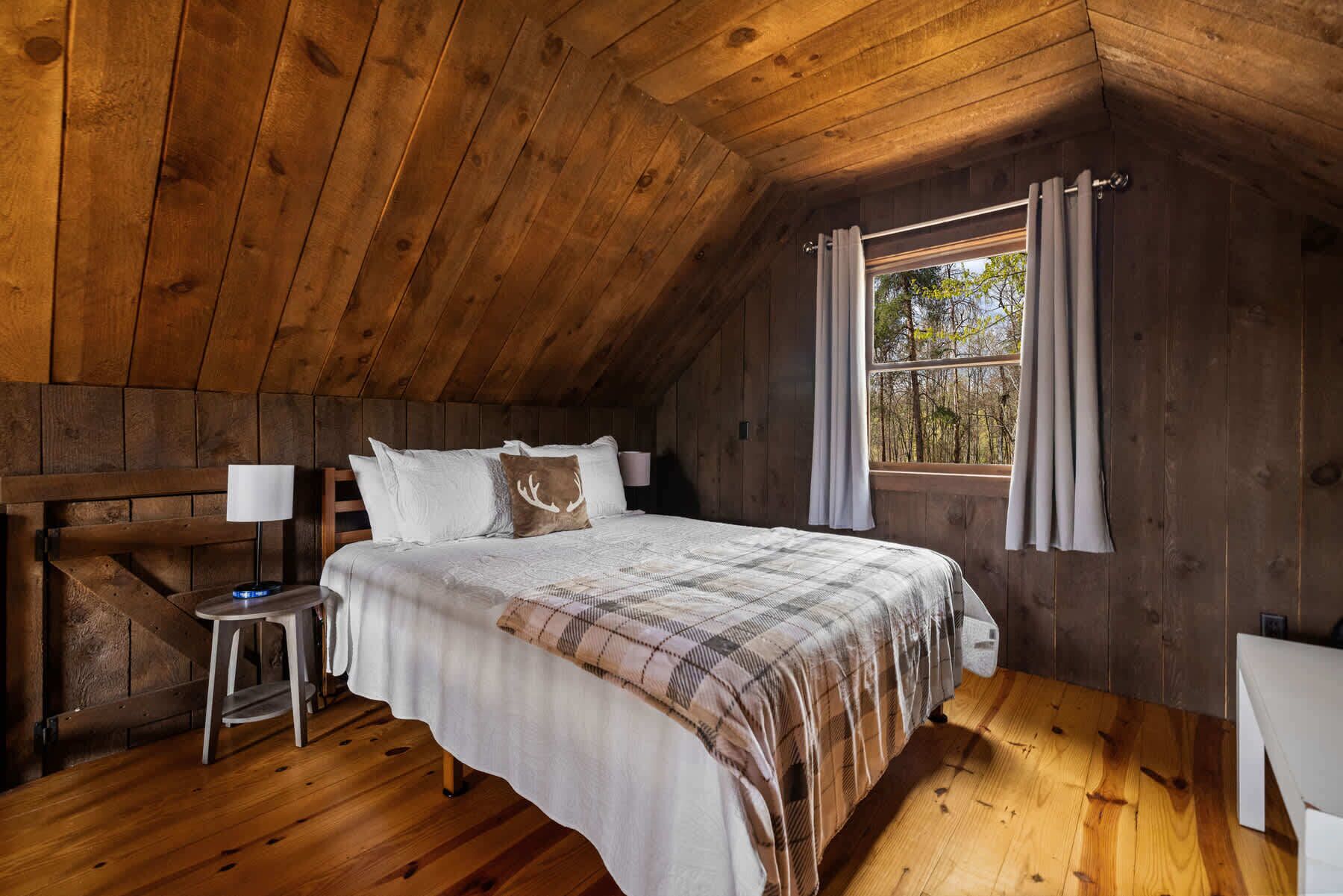 Una camera da letto mansardata accogliente, con soffitto in legno, un letto grande con copripiumino a quadri, un comodino con una lampada e una finestra con tende.