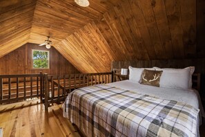 A cozy attic bedroom with a wooden sloped ceiling, a bed with a plaid bedspread, a window with a view of trees, and a wooden railing.