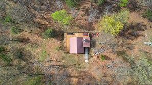 An aerial view of a single-story house with a red roof, surrounded by a wooded area.