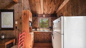 Una cocina rústica con paredes de madera, un refrigerador blanco, un banco de madera y una ventana con vistas a árboles.