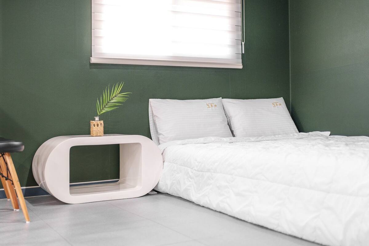 A bedroom with a bed, a bedside table, a small plant, and a window.