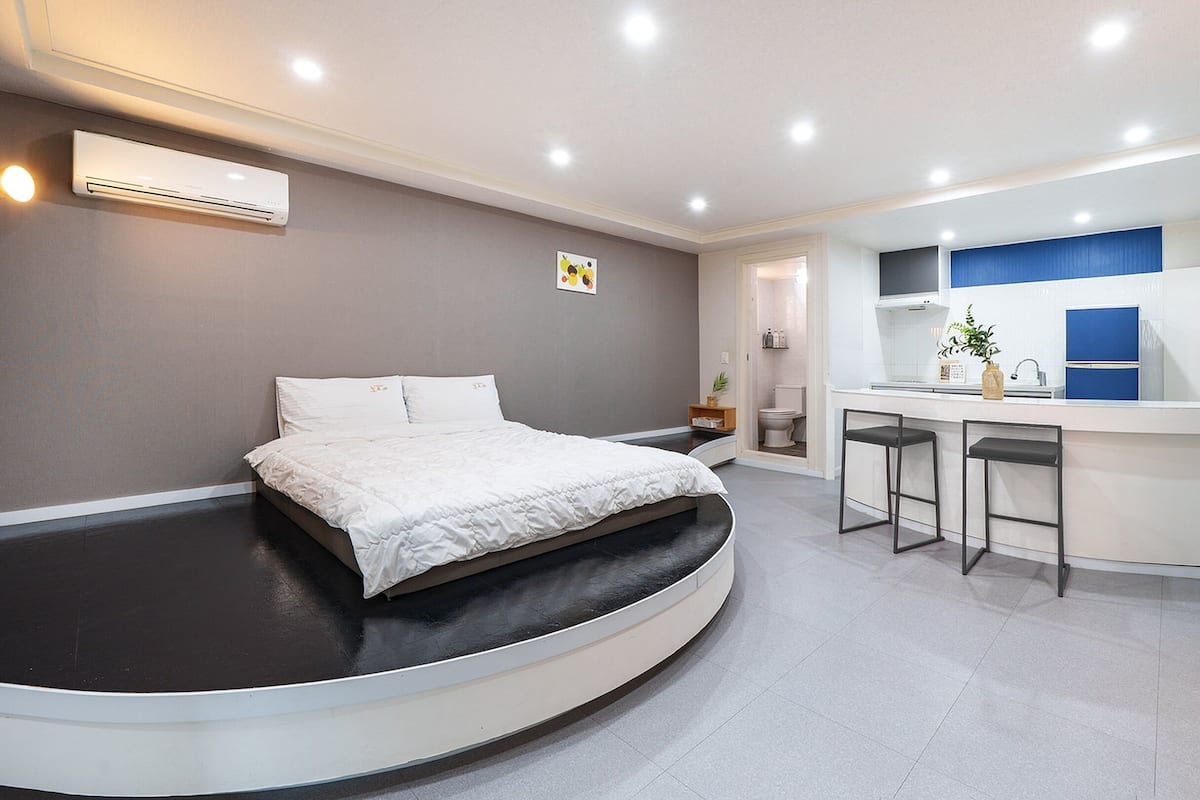 A modern bedroom with a curved bed, a small sitting area with stools, and a bathroom visible through an open door.