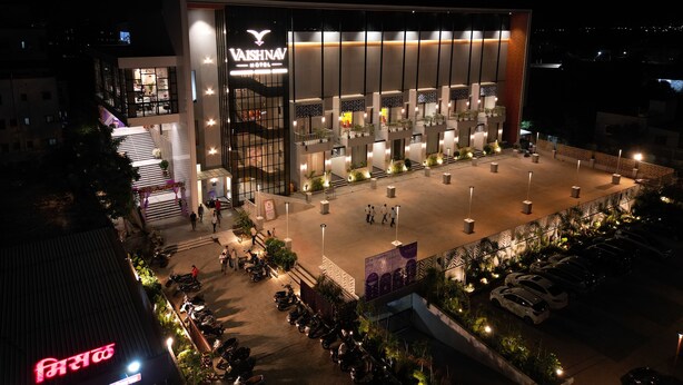 A modern hotel with a prominent sign, multiple balconies, and a spacious outdoor area with plants and seating. - Vaishnav Hotel (Latur)
