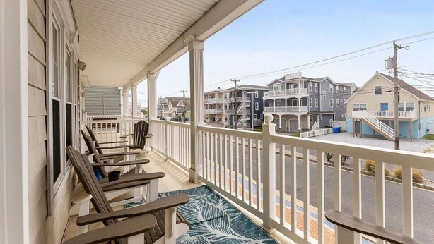A covered porch with wooden chairs and a patterned rug, overlooking a residential street with houses. - Modern 3 Bedroom Condo in a Prime Location (Wildwood)