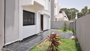A modern residential building with a clean, white exterior, a tiled patio, and a small garden area with a palm plant.