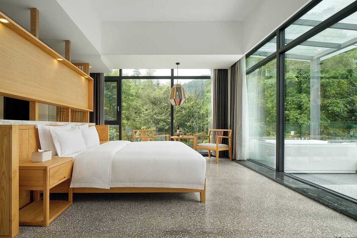 A modern bedroom with a large bed, wooden headboard, and a view of a lush garden through large windows.