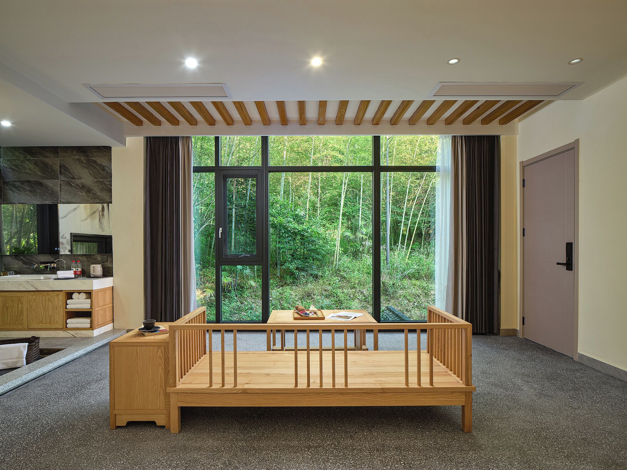A bedroom with a wooden bed, a large window with a forest view, a kitchen area, and a door.