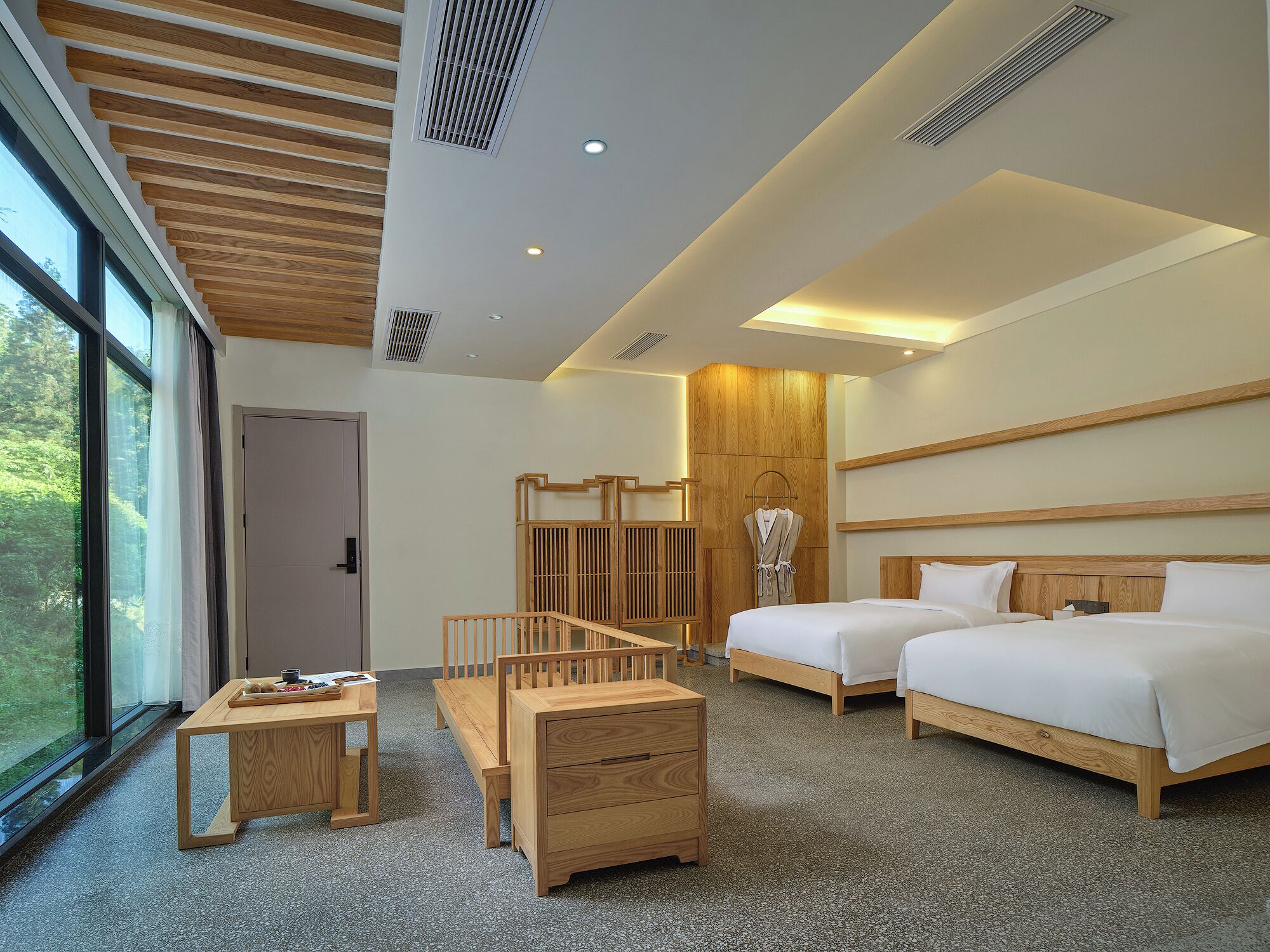 A modern hotel room with a large window, two beds, a wooden desk, and a small table.