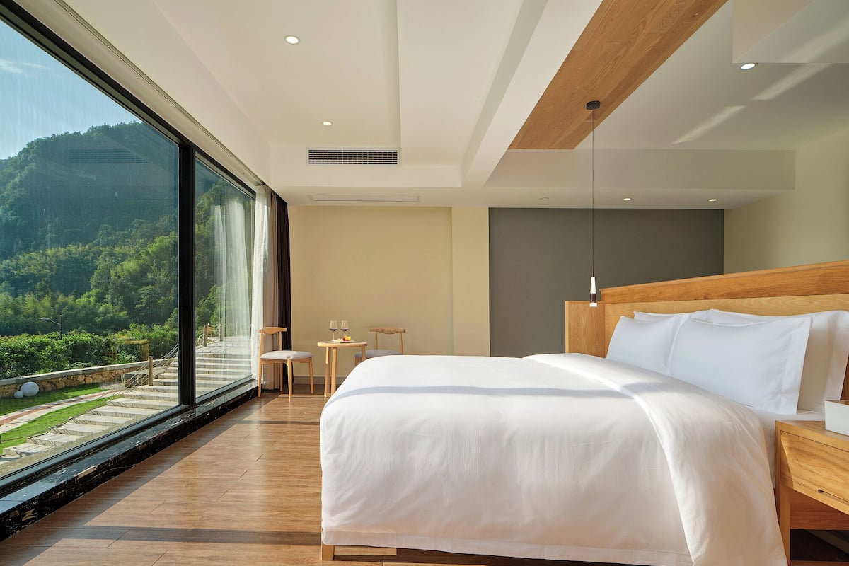 A modern hotel room with a large bed, a wooden headboard, and a view of a lush garden through floor-to-ceiling windows.