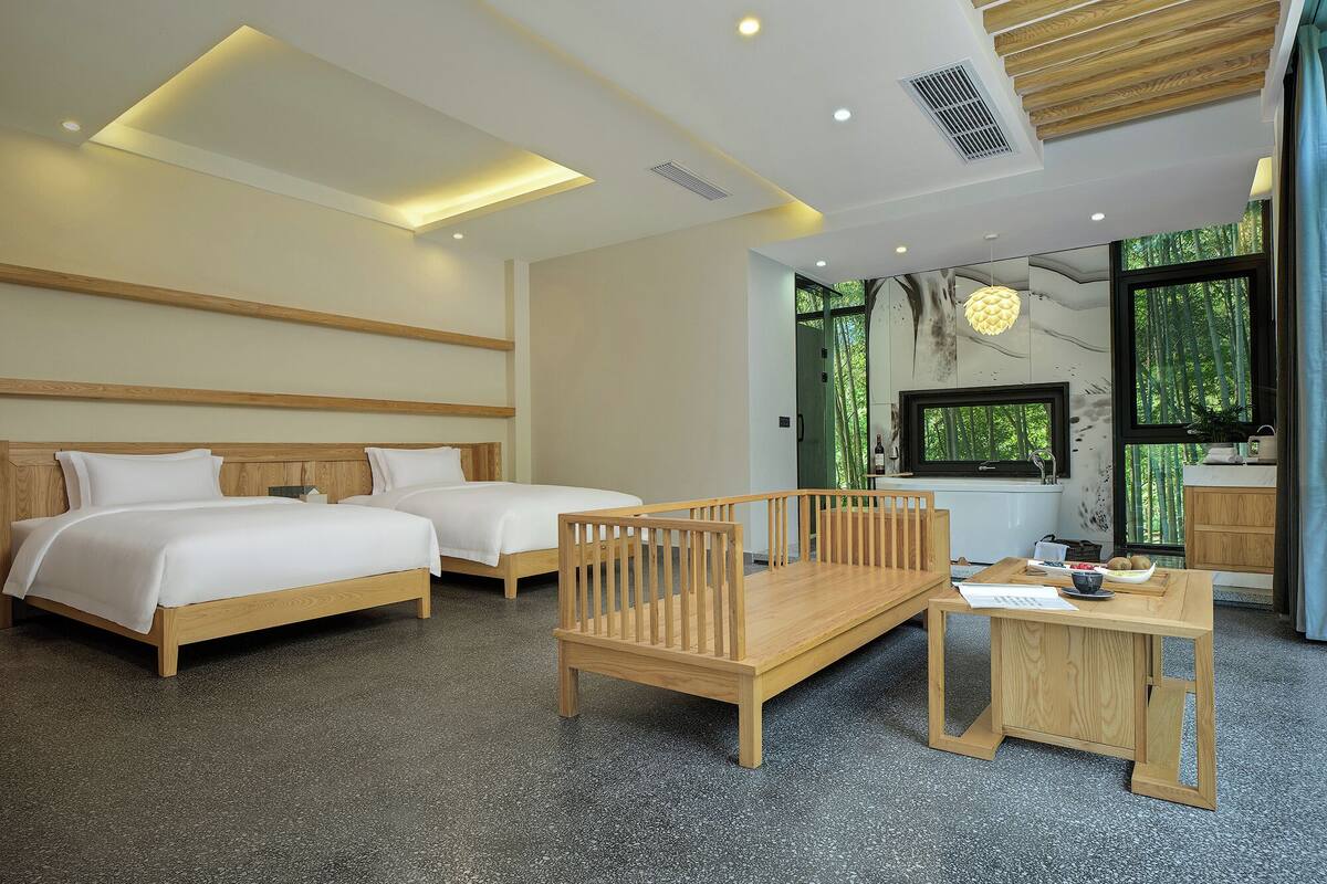A modern hotel room with two beds, a wooden bench, a small table, and a large window offering a view of trees.