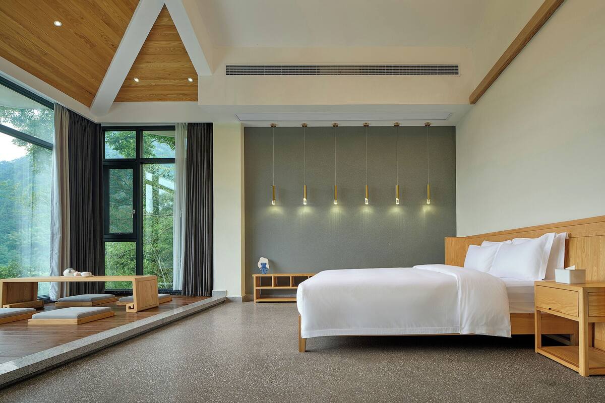 A modern bedroom with a large bed, wooden headboard, and a view of greenery through large windows.